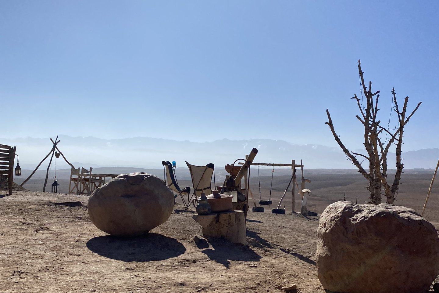 Camp dans le désert d'Agafay : sol de pierres et de gravier, gros blocs de roche, tapis berbère et chaises basses face à la ligne de l'Atlas